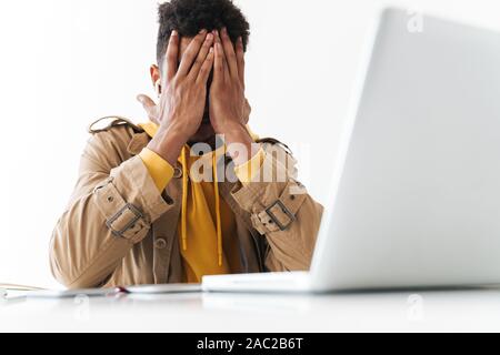 Image of upset african american man grabbing his face while working on laptop computer in white office Stock Photo