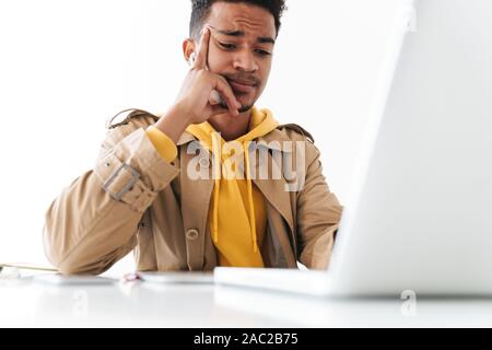 Image of young african american man thinking while working on laptop computer in white office Stock Photo