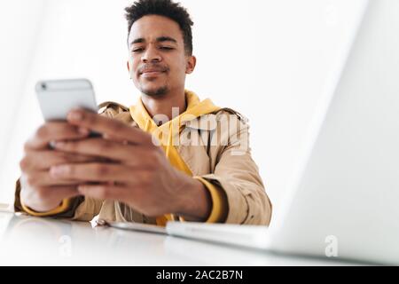 Image of focused african american guy using cellphone and laptop computer while sitting at table in white office Stock Photo