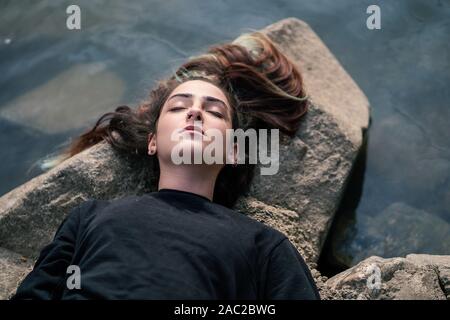 Girl lying and dreaming on rock near river bank. Relaxing in nature. Young woman sleeping on stone over water. Mindset reset