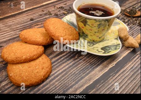 A Cup of black tea, tea leaves, pieces of brown sugar, oatmeal cookies on a wooden background. Close up Stock Photo