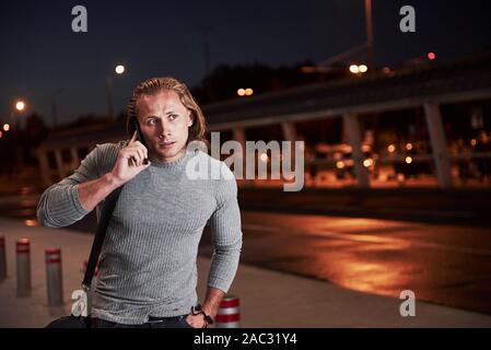 Having deal with some things. Young stylish man walking through the night street near the road and having a conversation on the phone Stock Photo