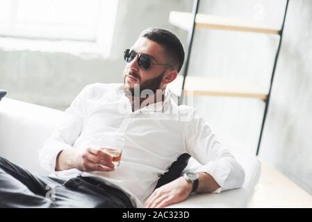 Thoughtful look. Photo of young bearded businessman in sunglasses and whiskey in the hand sit on the white sofa Stock Photo