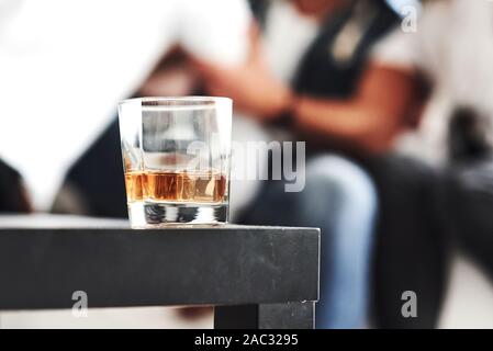 Close up focused photo of glass with whiskey standing on the black table with blurred people on the background Stock Photo