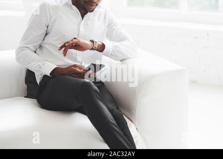 Cropped photo. Checking the time. Young afro american guy in office wear sitting on the white sofa Stock Photo