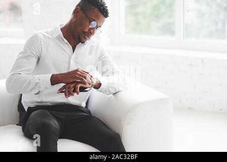 Checking the time. Young afro american guy in office wear sitting on the white sofa Stock Photo