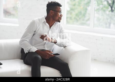 Looking to the side. Young afro american guy in office wear sitting on the white sofa Stock Photo