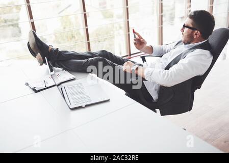 Young businessman having a rest while sitting in chair and put legs on the table in the clean room. Watching through the windows Stock Photo