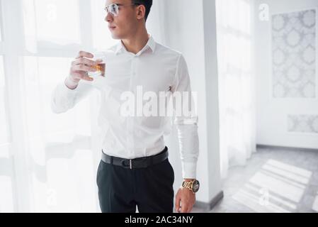 Thoughtful view. Gorgeous man in eyeglasses stand indoor and drink alcohol Stock Photo