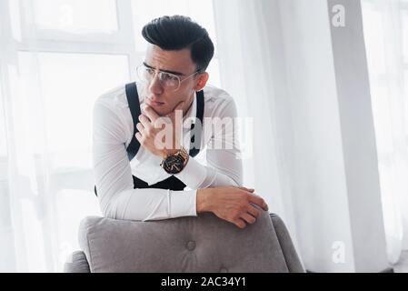 Thinks about something. Young good looking man in classical clothes and eyewears leaning on the chair while standing against white windows Stock Photo