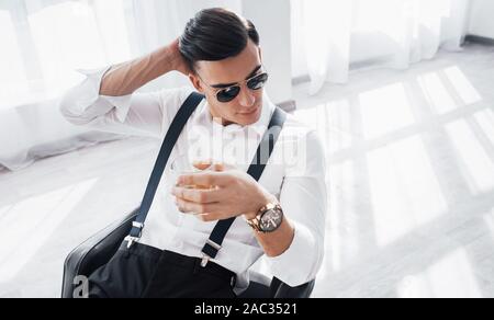 You can see the big veins on the right arm. Young stylish businessman sitting on a black chair with glass of whiskey Stock Photo