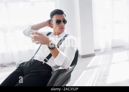 Joyful mood. Young stylish businessman sitting on a black chair with glass of whiskey Stock Photo