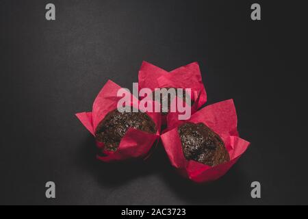Three brown baked chocolate muffins rolled in pink paper and placed on matte black background with copy space Stock Photo
