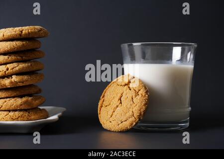 A gingerbread cookie leans on a glass of cold milk next to a stack of cookies on a white plate with a black background; copy space Stock Photo