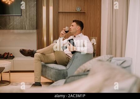 Young man sits in a luxury apartment, drinking red wine and holding a digital tablet Stock Photo