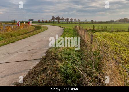 Bicycle lane beside a road with dangerous curve marking between farmland with fences and trees in the background, otonal day on a cloudy day Stock Photo