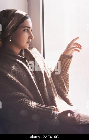 Young Muslim woman sits on window sill and looking away. Depressed middle-eastern female near the window at home. Lonely cheerless girl feeling stress Stock Photo