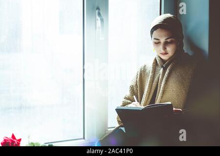 Young Muslim woman sits on the window sill. Middle-eastern writer woman sitting thoughtfully near the window and writing her story on the notepad. Ira Stock Photo