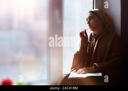 Young Muslim woman sits on the window sill. Middle-eastern writer woman sitting thoughtfully near the window and writing her story on the notepad. Ira Stock Photo