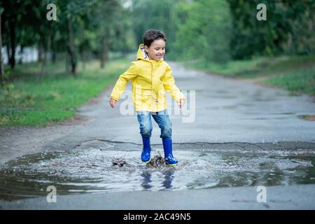 A wet child is jumping in a puddle. Fun on the street. Tempering in summer. Splashes, drops of water, outdoor. waterproof boots jump in puddle and mud Stock Photo