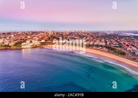 Smooth waters of Pacific ocean surfing on famous Sydney Bondi beach at sunrise in aerial view towards distant city CBD towers and Sydney harbour bridg