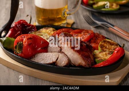 Close up of grilled meat on cast Iron pan with grilled vegetables and beer on rustic wooden table. Stock Photo