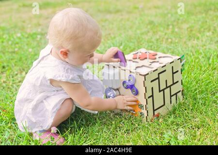 Cute little girl is playing with busiboard outdoors on green grass. Educational toy for toddlers. girl opened door to cube of board Stock Photo