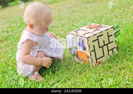 Cute little girl is playing with busiboard outdoors on green grass. Educational toy for toddlers. girl opened door to cube of board Stock Photo