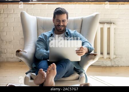 Smiling relaxed young man sit on armchair using laptop laughing Stock Photo