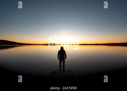 Lonely man's silhouette stands on big rock at sunset lake and looks on mountains Stock Photo