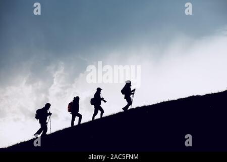 Group of four hikers silhouettes are going uphill in mountains against cloudy sky Stock Photo