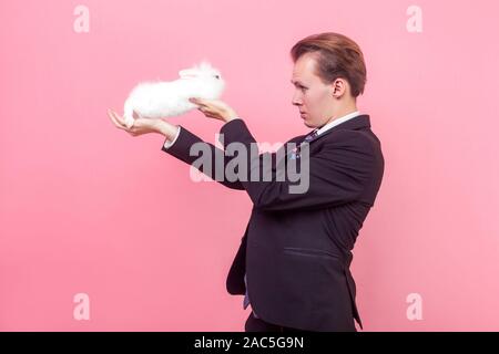 Side view of serious young man in elegant suit and with stylish hairdo holding white fluffy rabbit on raised hands and looking at bunny with curiosity Stock Photo