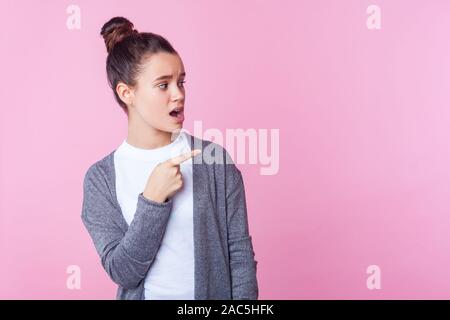 Wow, advertise here! Portrait of wondered teenage girl with bun hairstyle in casual clothes looking with open mouth in surprise, pointing to the side. Stock Photo