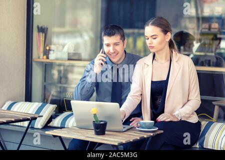 Smiling business people using laptop at cafe terrace while talking on phone – remote working Stock Photo