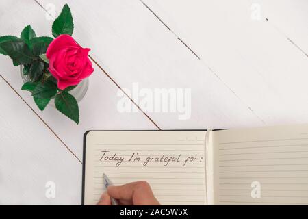 Woman hand writing in a gratitude journal on white wooden table background with pink rose decoration Stock Photo