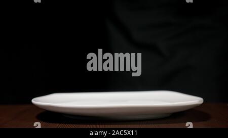 White plate of square shape. The dishes are on a black background on a brown table. Side view. Stock Photo