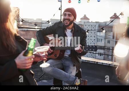 Photo of young bearded attractive guitarist that sings for his friends on the rooftop Stock Photo