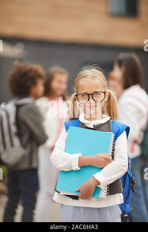 Portrait of blond schoolgirl in eyeglasses holding textbooks and looking at camera while standing outdoors with her classmates in the background Stock Photo