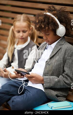 African schoolboy in headphones using his mobile phone and listening to music while sitting on the bench together with girl outdoors Stock Photo