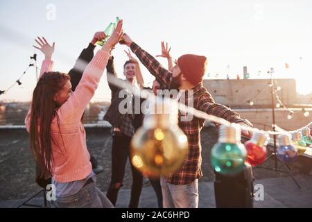 Blurred light bulbs in the front of camera. Holidays on the rooftop. Cheerful group of friends raised their hands up with alcohol Stock Photo