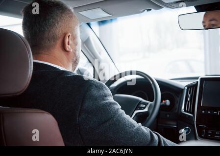 Reflection of face in the front mirror. View from behind of senior businessman in official clothes driving a modern new car Stock Photo