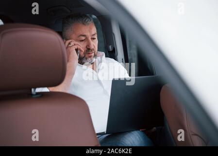 Talking on the phone. Working on a back of car using silver colored laptop. Senior businessman Stock Photo
