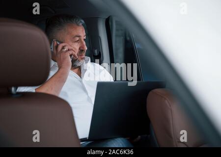 Having business call while sitting at the back of car with silver colored laptop Stock Photo