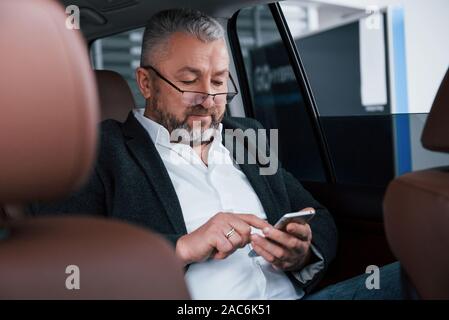Contacting with colleagues. Using mobile phone. Businessman in the eyeglasses sits in the back of the modern car and have some deals Stock Photo