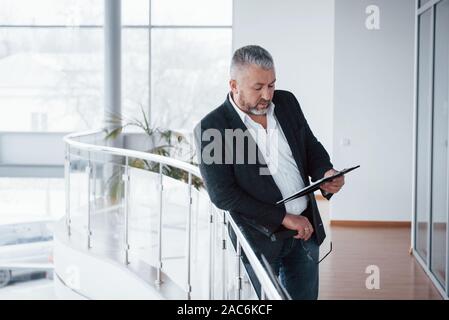 Big windows provide excellent lighting. Photo of senior businessman in the spacious room with plants behind. Holding and reading documents Stock Photo