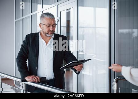 Hand of employee is about to take that papers. Photo of senior businessman in the spacious room with plants behind. Holding and reading documents Stock Photo