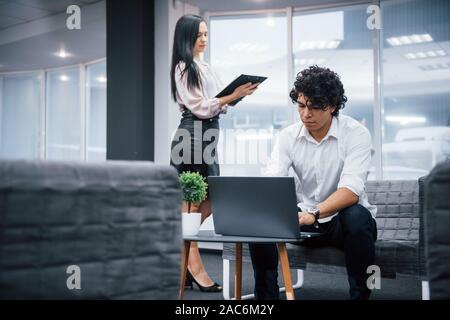 Freelance work in the office with windows behind. Two people is on their job. Guy using silver laptop. Girl read document Stock Photo