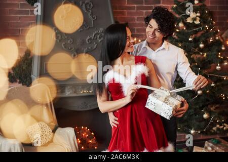 I guess it's time for open it. Beautiful couple celebrating New year in the decorated room with Christmas tree and fireplace behind Stock Photo