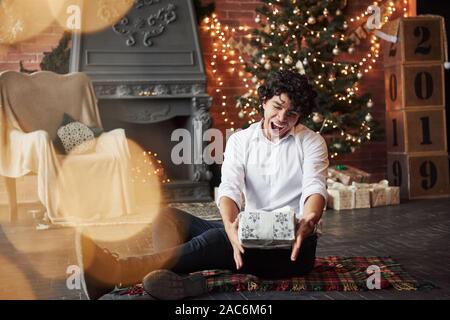 Finally it's holidays. Guy is satisfied. Young man sitting on the floor of beautiful decorated room and holding white gift box in the New year time Stock Photo