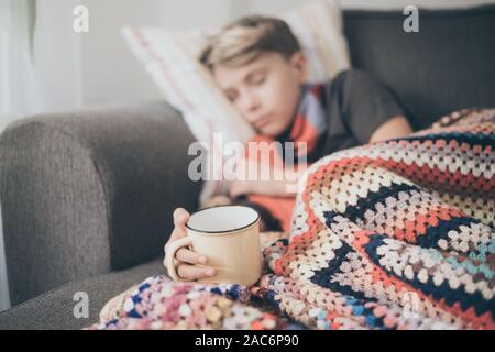 Sick boy sleeping with woolen blanket, hot water bottle and a mug. Sad teen with the flu rests alone at home in a cold winter day. Child with seasonal Stock Photo